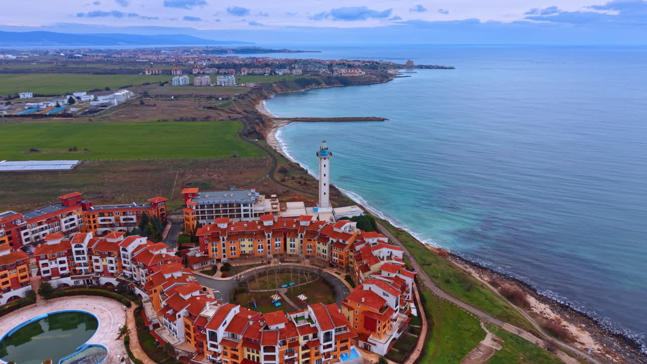 Vista aerea di un resort costiero con tetti rossi e faro. Architettura circolare di un complesso residenziale vicino alla costa turchese con faro e campi verdi.