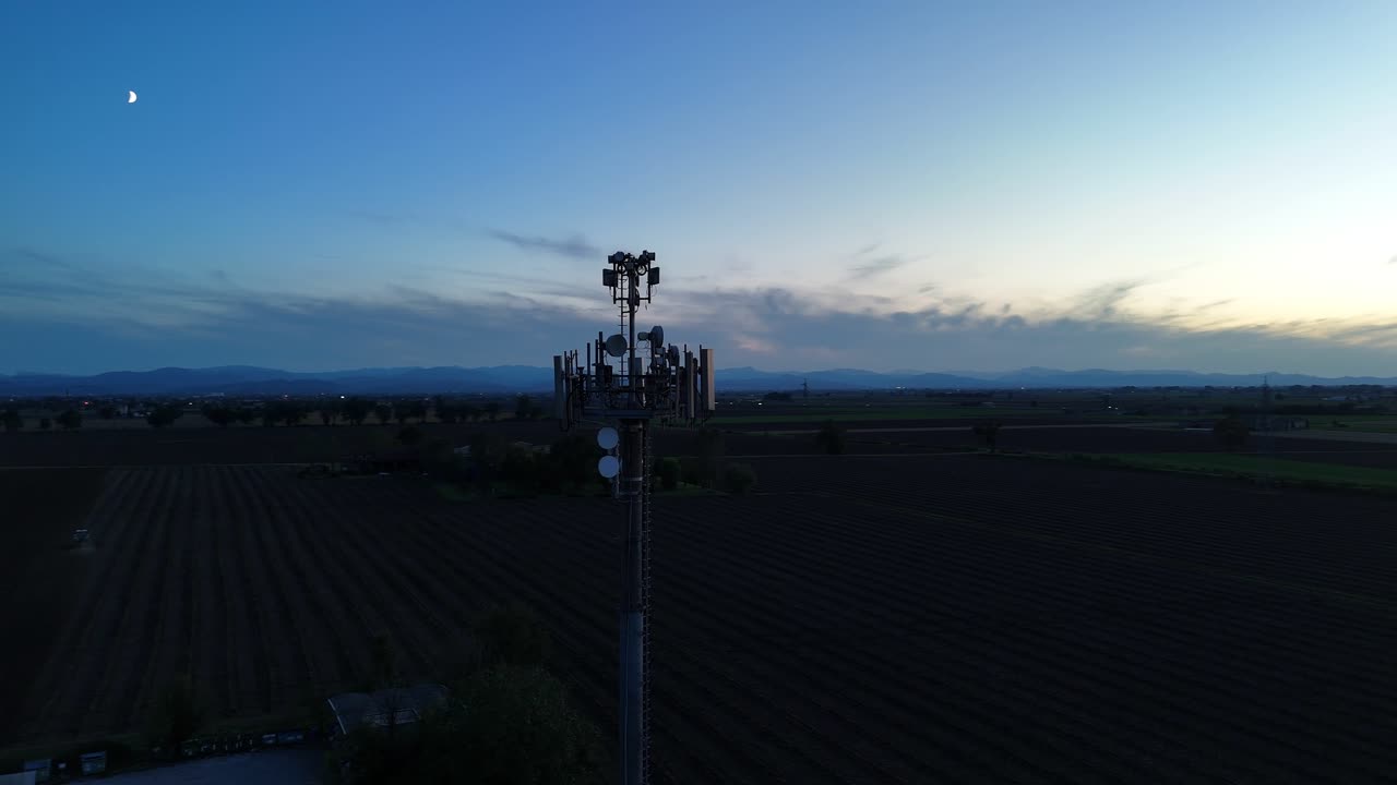 Filmato con drone che riprende una moderna torre di telecomunicazioni con antenne contro un cielo crepuscolare su campi rurali italiani, evidenziando l'infrastruttura di connettività.