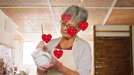 animation of heart icons over caucasian female worker painting pottery