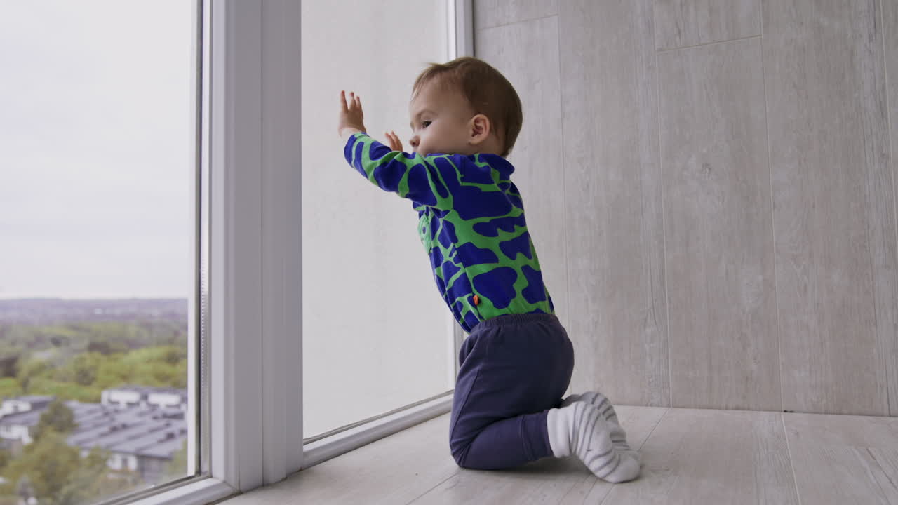 Caucasian baby boy playing on the floor near the big window. Toddler banging by the window with his hands and than distracts on something else.