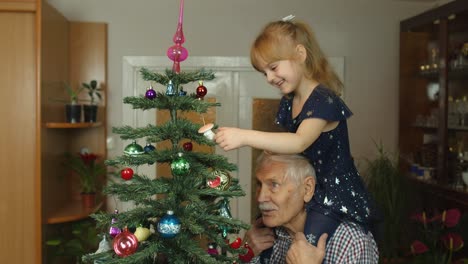 Girl-kid-with-senior-grandpa-decorating-artificial-Christmas-tree-with-ornaments-and-toys-at-home