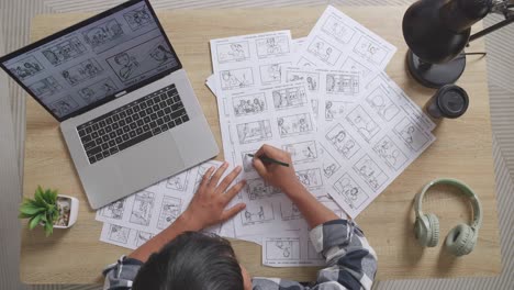 top view zoom out shot of asian male artist drawing storyboard for the film on the table with a laptop and headphones in the studio