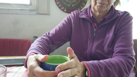 una anciana caucásica se calienta las manos sosteniendo una taza de té caliente en el interior