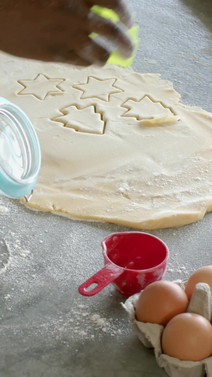 Vertical video: Reaching maroon arm pressing green cutter into dough, making tree cookies with eggs
