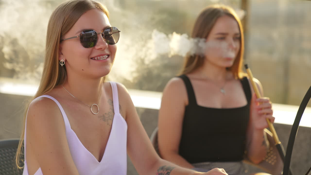 Friends having relaxing time together in a café outdoors. Summer leisure communicating and smoking hookah. Sunny blurred backdrop.