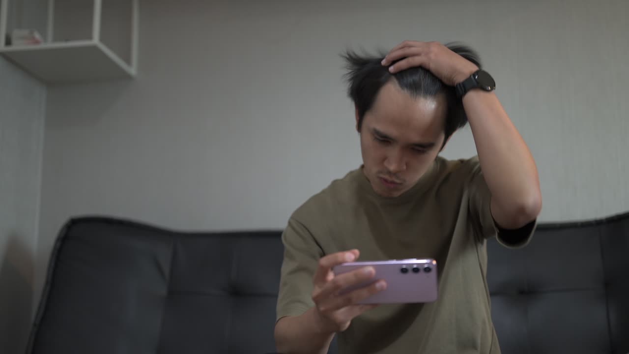 Handsome young Asian Thai man sitting at sofa indoors while playing games with smartphone