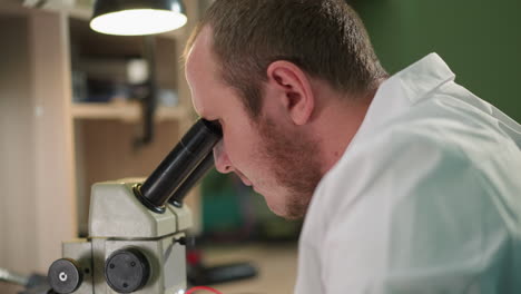 una vista cercana de un técnico en una bata de laboratorio blanca observando cuidadosamente algo bajo un microscopio con un alambre rojo en la mano en un laboratorio, el espacio de trabajo tiene una lámpara de escritorio