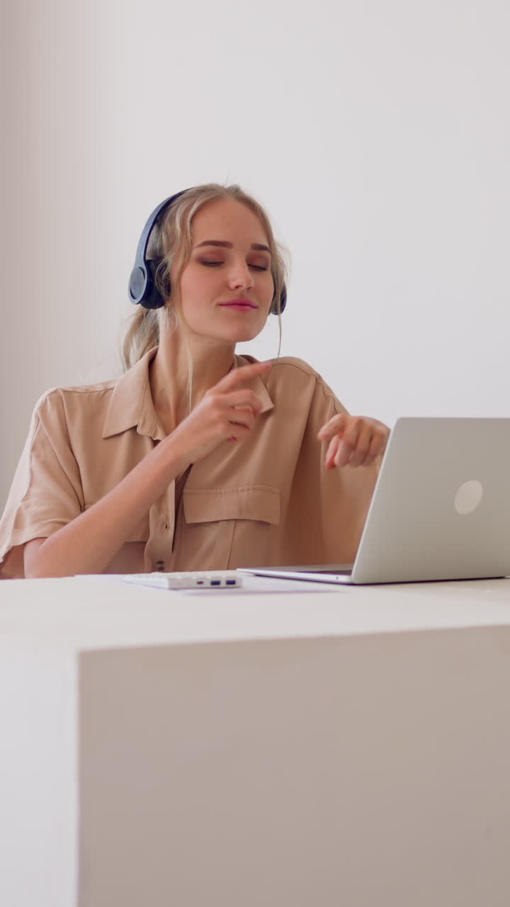 Young woman with headphones moves to rhythm of dynamic music sitting at white desk with laptop in comfortable light office slow motion