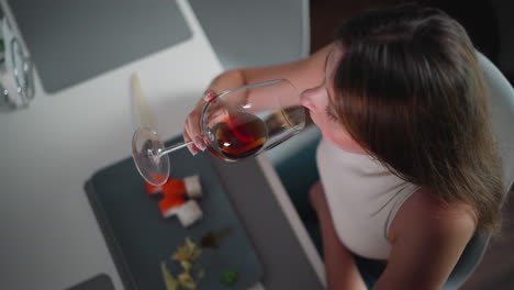 business lady in sleeveless outfit sips red wine at dining table with plate of sushi in front, top view angle shows relaxed dining experience in stylish modern setting with natural indoor light