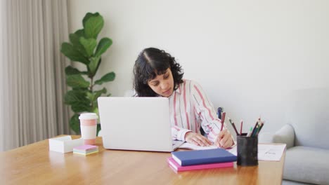 Biracial-woman-sitting-at-desk-using-laptop-and-writing-in-living-room