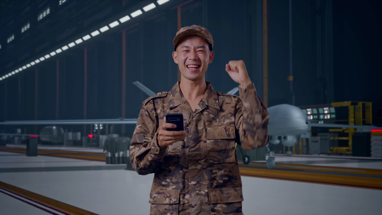 Soldato asiatico che guarda lo smartphone, poi urla "gol" festeggiando mentre è in piedi con un moderno drone militare in un hangar di manutenzione.