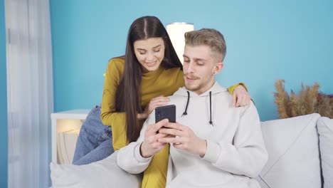 행복한 젊은 부부가 집에서  ⁇ 드폰을 보고, 온라인 쇼핑을 하고, 소셜 미디어 게시물을 확인하고 있습니다.