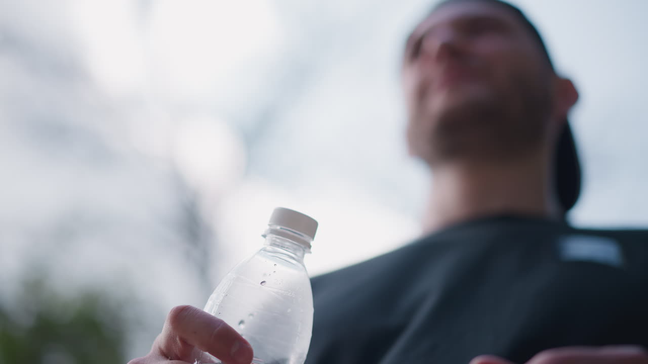 Athlete Drinks Water, Individual Rehydrates With Bottled Water During Training Outside In Nature, Athlete Takes Quick Drink From Plastic Bottle While Training Outdoors In Front Of Cloudy Sky