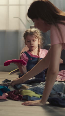 upset little girl looks at mother gathering clothes scattered on floor near suitcase in living room. woman cleans up mess after daughter play