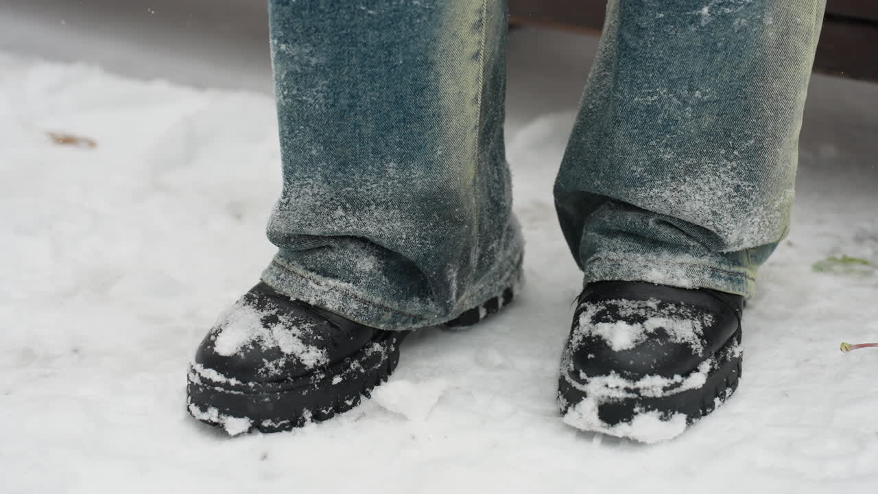 primer plano de piernas en botas negras cubiertas de nieve tocando rítmicamente en el suelo blanco, vaqueros de vaqueros salpicados de helada, capturando un momento invernal de movimiento sutil en medio de un entorno sereno nevado