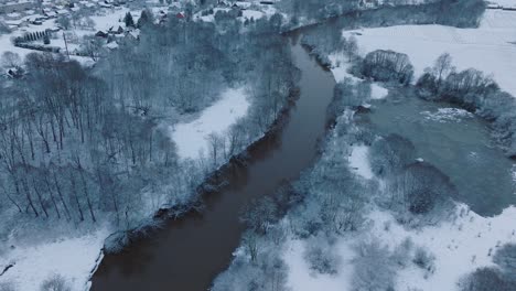 Río-Sinuoso-A-Través-De-Un-Bosque-Cubierto-De-Nieve-En-El-Valle-Del-Río-Abava-Cerca-Del-Pueblo-De-Renda,-Vista-Aérea,-Movimiento-Hacia-Adelante,-Inclinación-Hacia-Arriba