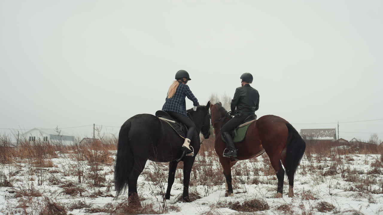 Due cavalieri a cavallo in un campo innevato che si scambiano saluti e aggiustano le redini, caschi e giacche invernali visibili, lezione cooperativa in tandem che enfatizza il lavoro di squadra, la fiducia e l'equilibrio in un contesto rurale