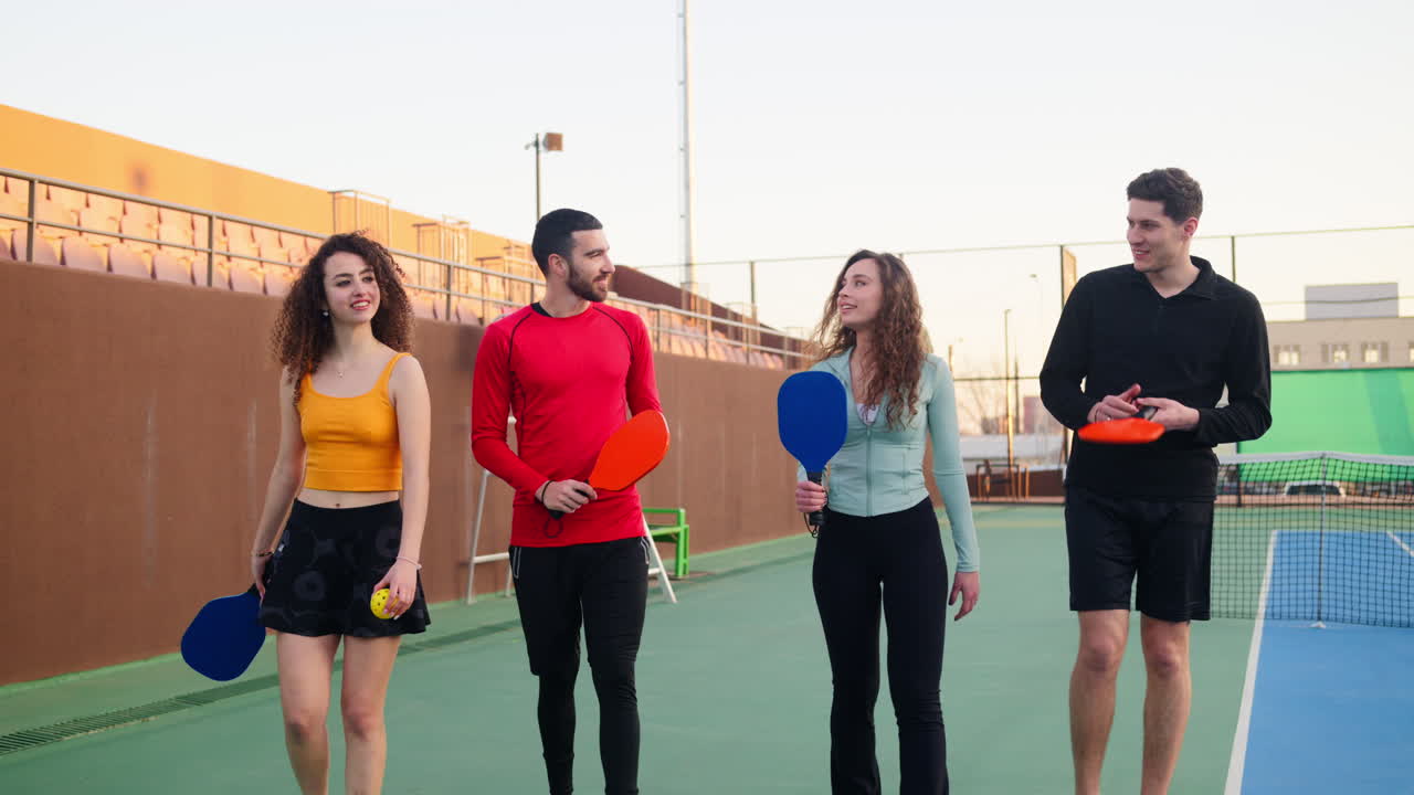 Two men and two women holding pickleball rackets walking, talking and smiling on a court. Group of friends