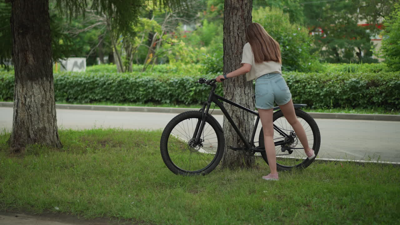 청바지를 입은 짧은 여성은 평화로운 공원 환경에서 나무에 기대어 자전거를 접근, 나무의 울창한 녹색 배경과 포장 길을 제거 준비