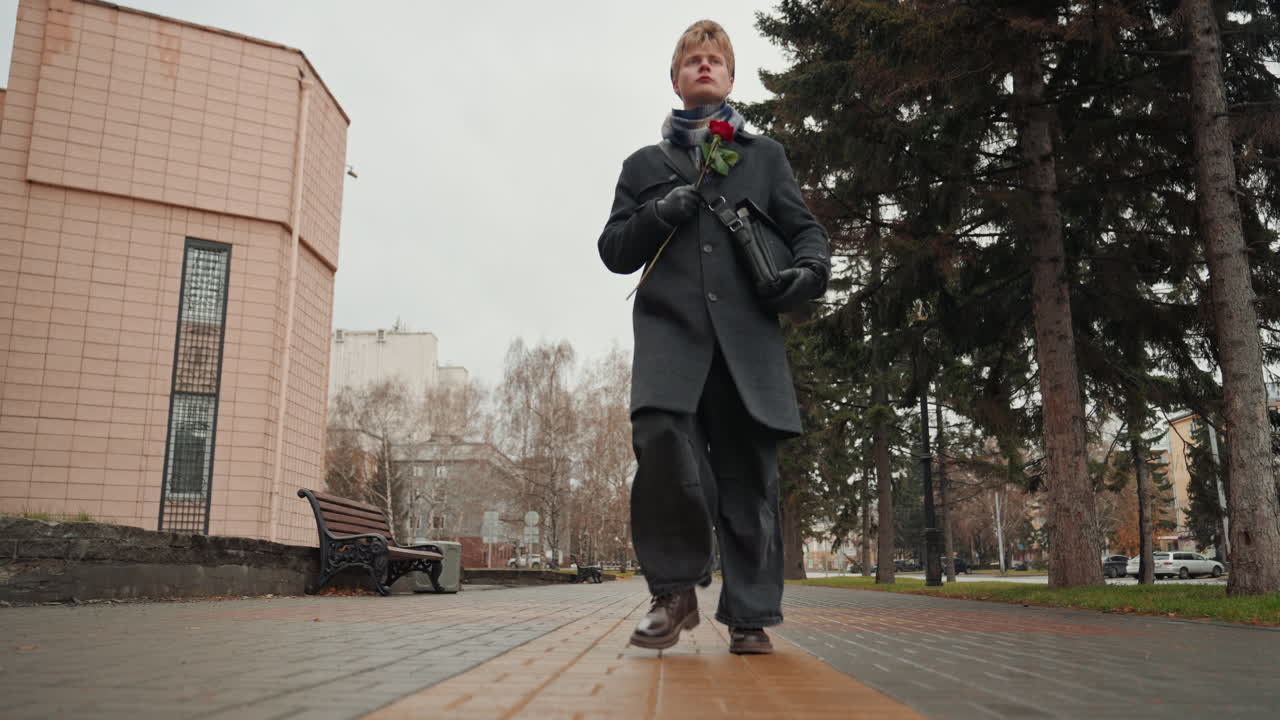 White man walking down city path holding rose, creator in long coat strolling past pine trees and brick building on wet pavement, lowangle cinematic shot emphasizing boots and steady steps, overcast