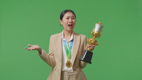 황금 메달을 가진 슈트를 입은 아시아 사업가 여성이 스튜디오의 녹색 스크린 배경에서 트로피를 의심스럽게보고 있습니다.