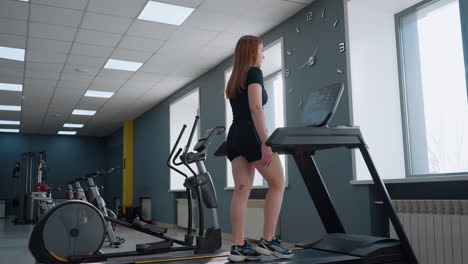 sport woman keeps fit walking on treadmill under sunlight through large window in modern gym with wall clock on gray wall row of exercise bikes and elliptical trainer visible in background