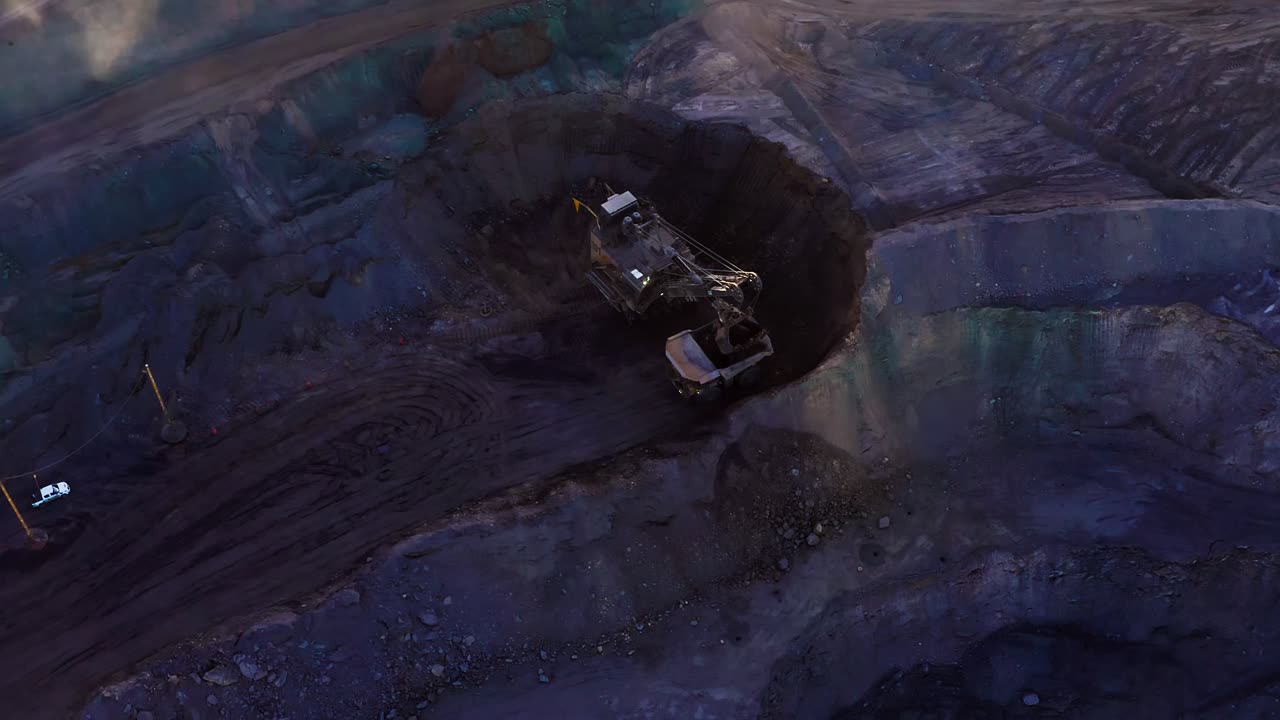 Drone orbits excavator filling dump truck at bottom of open gold pit mine, highlighting scale, teamwork, and mechanized extraction, and environmental degradation with water safety and pollution