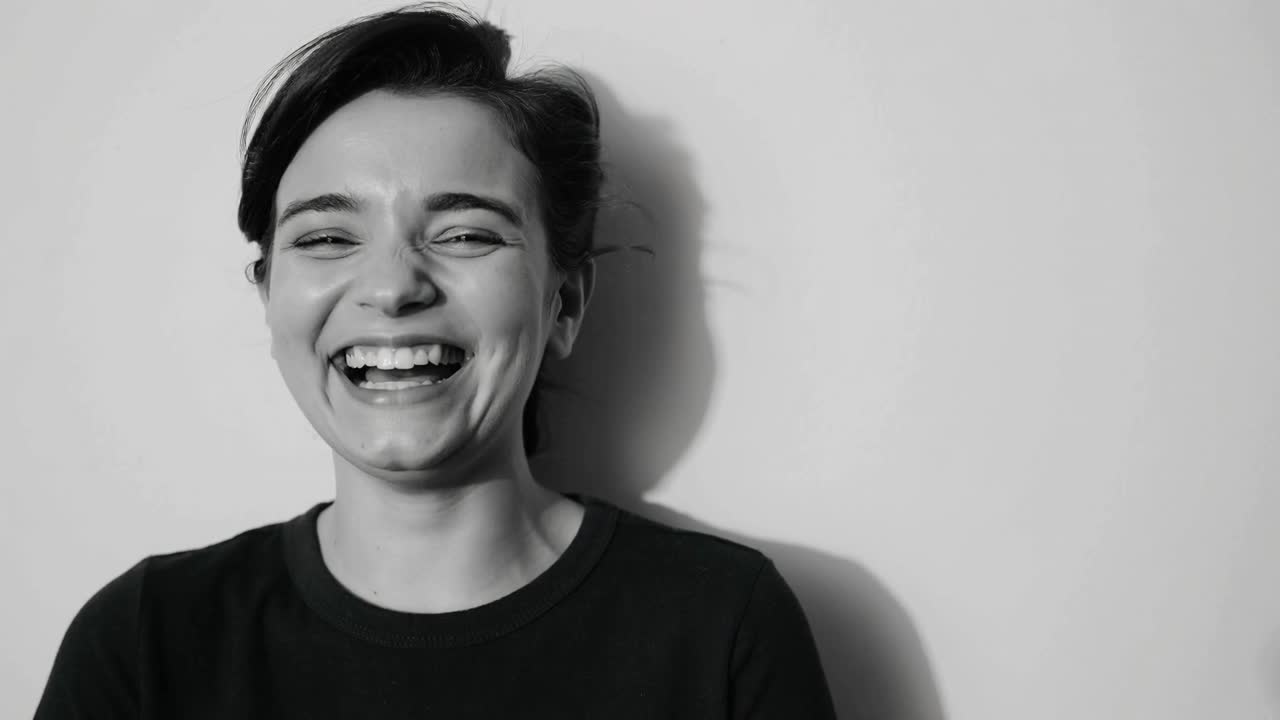 Una donna che da un sorriso passa a una risata dopo un segnale fuori campo, con una parete chiara, una maglietta scura e un'ombra in movimento sullo sfondo.