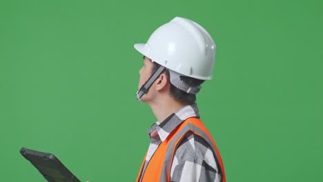 close up side view of asian male engineer with safety helmet looking at the tablet in his hand and looking around while standing in the green screen background studio