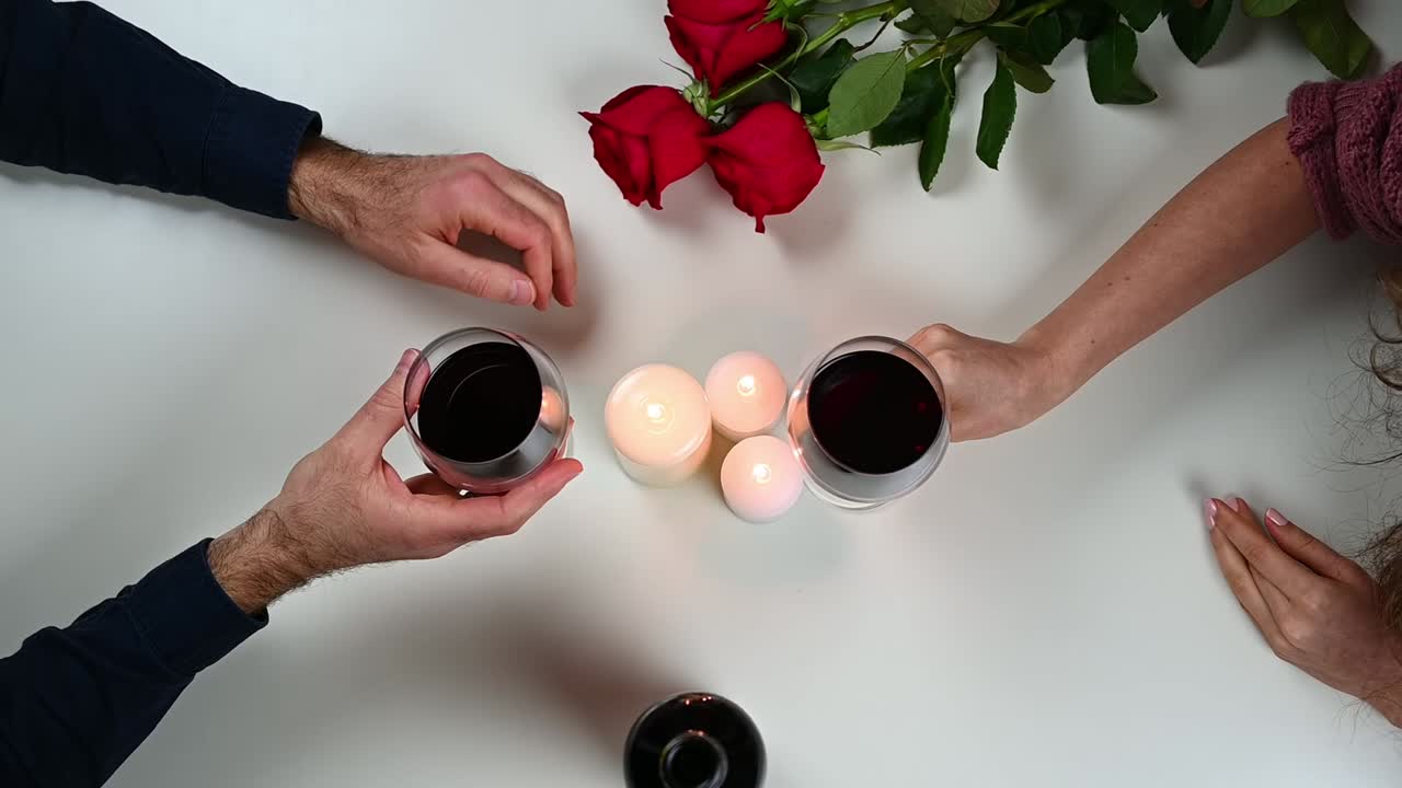 Woman and man are holding hands and drinking wine among red roses and candles. White table. Valentines day in love. Relationships and feelings concept. Overhead shot. Slow motion