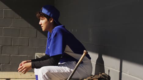 Nervöse-Gemischtrassige-Baseballspielerin,-Die-Auf-Einer-Bank-In-Der-Sonne-Sitzt-Und-Mit-Einem-Baseballball-Wartet