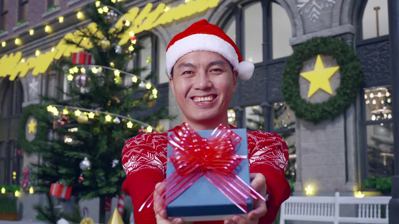 산타 의상을 입은 아시아인 남자가 크리스마스 밤에 서 있는 동안 선물을 보여주고 카메라에 미소 짓고 있습니다.