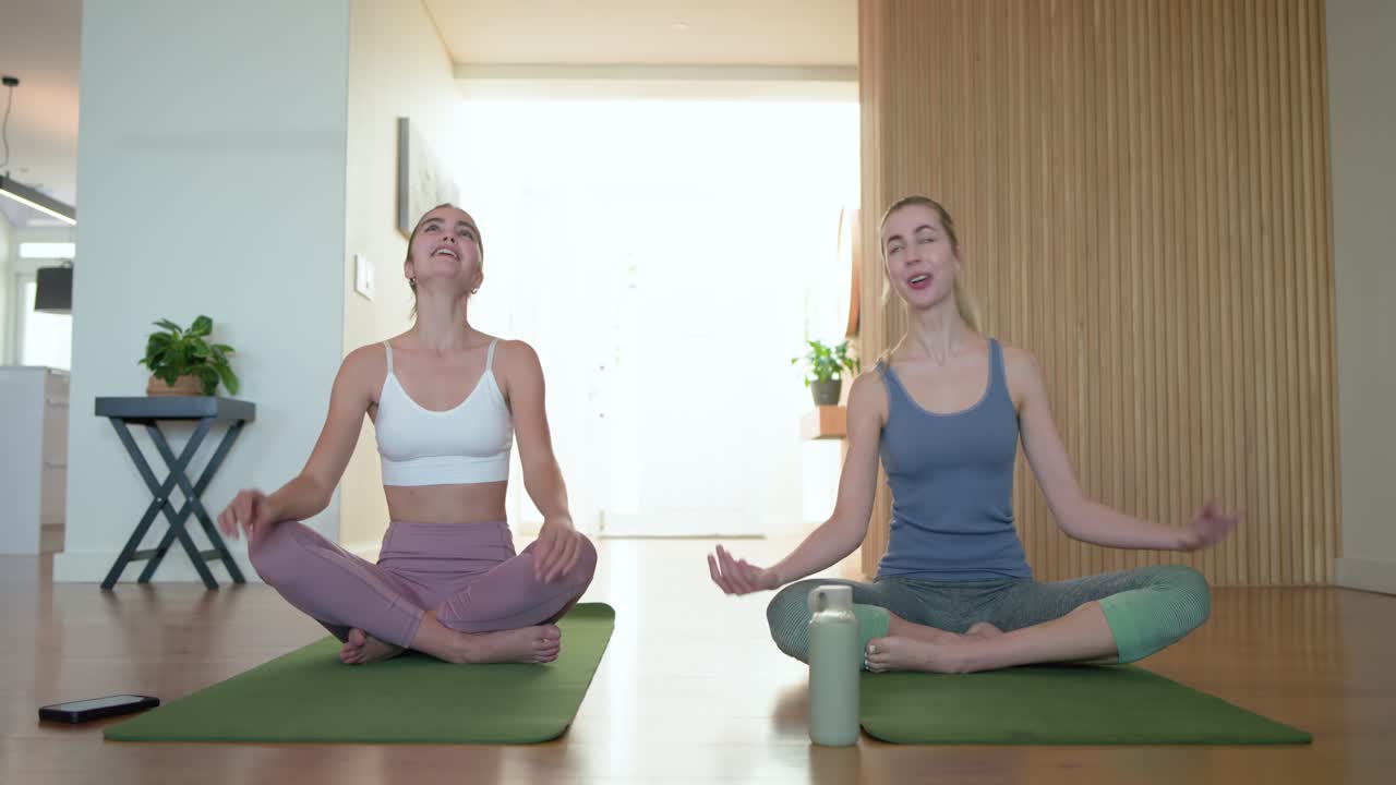 Due donne che completano la meditazione e aprono gli occhi, gesticolando su tappetini da yoga verdi in uno studio domestico