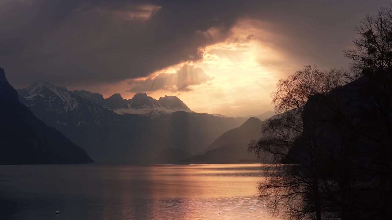Intense golden sunset rays bursting through storm clouds over alpine lake with snowy peaks and tree silhouette Switzerland