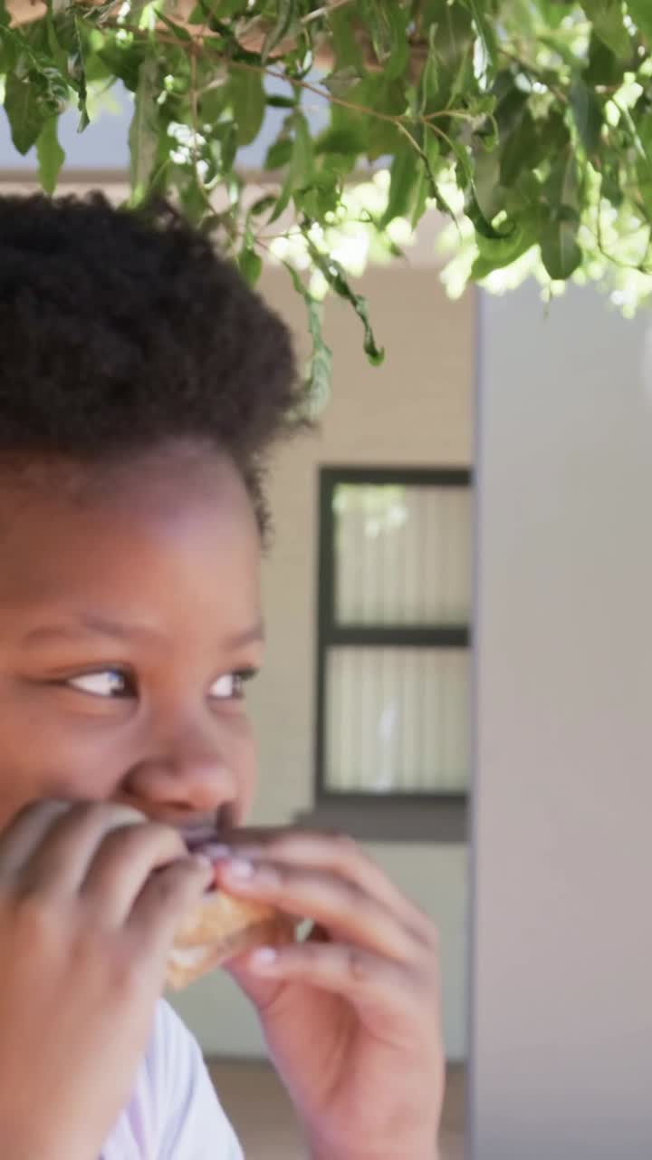 Vertical video: Holding wrapped bun, girl in pink top snacking by facade under leaves, copy space