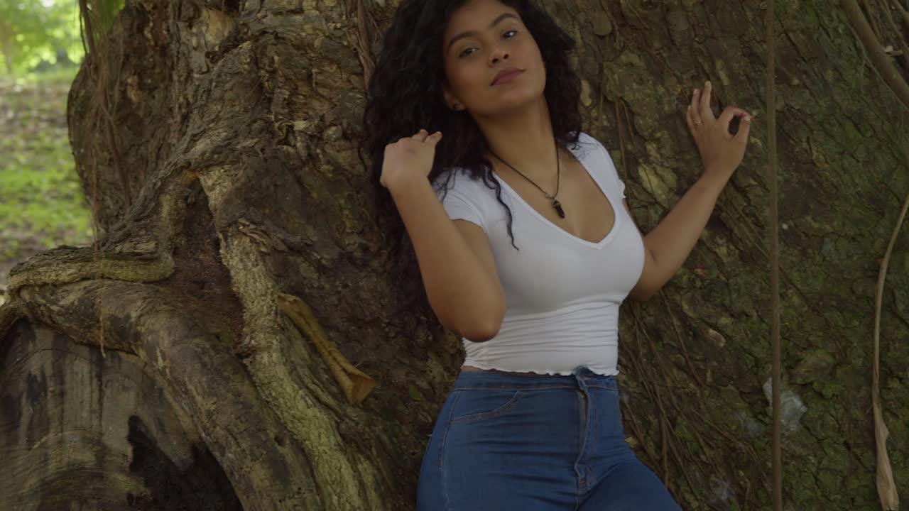 큰 나무 줄기에 공원에 서있는 섹시한 젊은 히스패닉 소녀