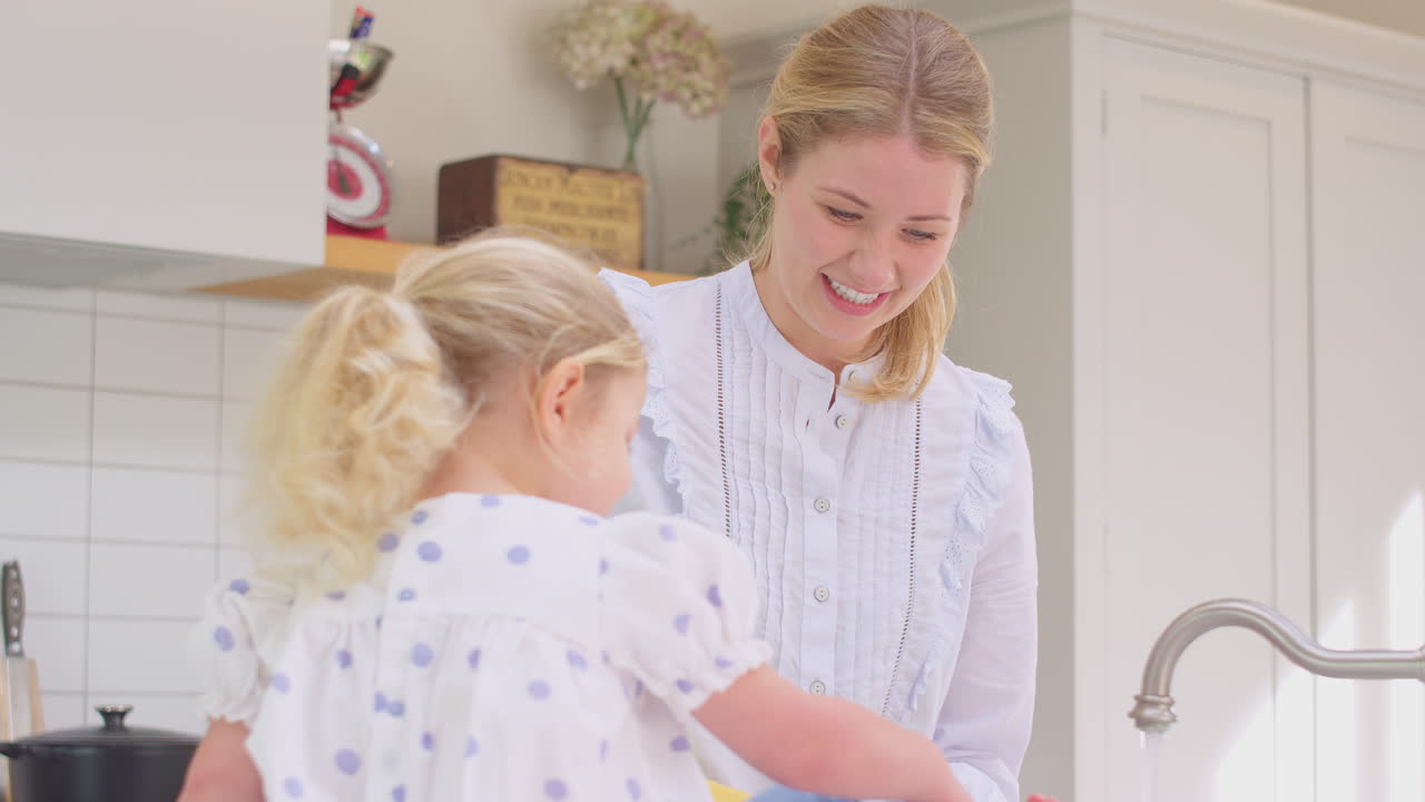 Mother wearing rubber gloves at home in kitchen with young daughter having fun and washing girl's feet as they do washing up at sink- shot in slow motion