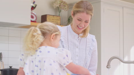 Mutter-Trägt-Gummihandschuhe-Zu-Hause-In-Der-Küche,-Während-Ihre-Kleine-Tochter-Spaß-Hat-Und-Mädchen-Die-Füße-Wäscht,-Während-Sie-Am-Waschbecken-Abwasch-Machen-–-Aufnahme-In-Zeitlupe