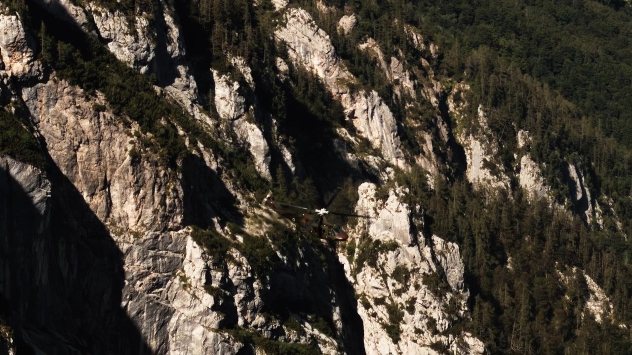 Camouflaged military helicopter flies past steep rocky mountains and dense forest, showing aviation and defense operations in rugged alpine terrain.