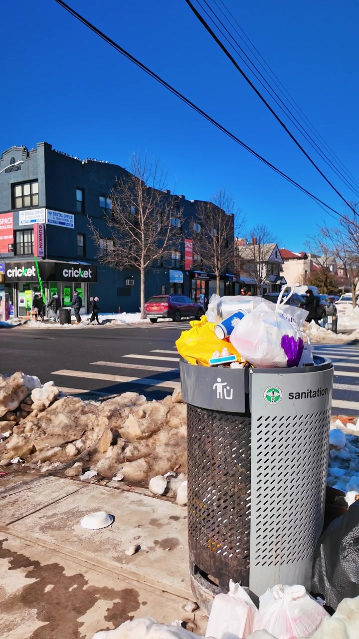 New York, USA, 5 February 2026: Overfilled public trash can on a snowy city sidewalk. Silver metal sanitation bin overflowing with garbage bags and litter on a sunny winter day in the city.