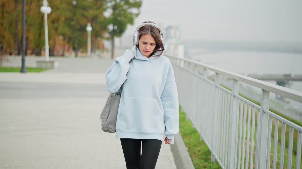 Young woman in blue hoodie thoughtfully listening to music, adjusting her bag while walking slowly along iron rail, enjoying outdoor stroll, calm and serene moment in nature