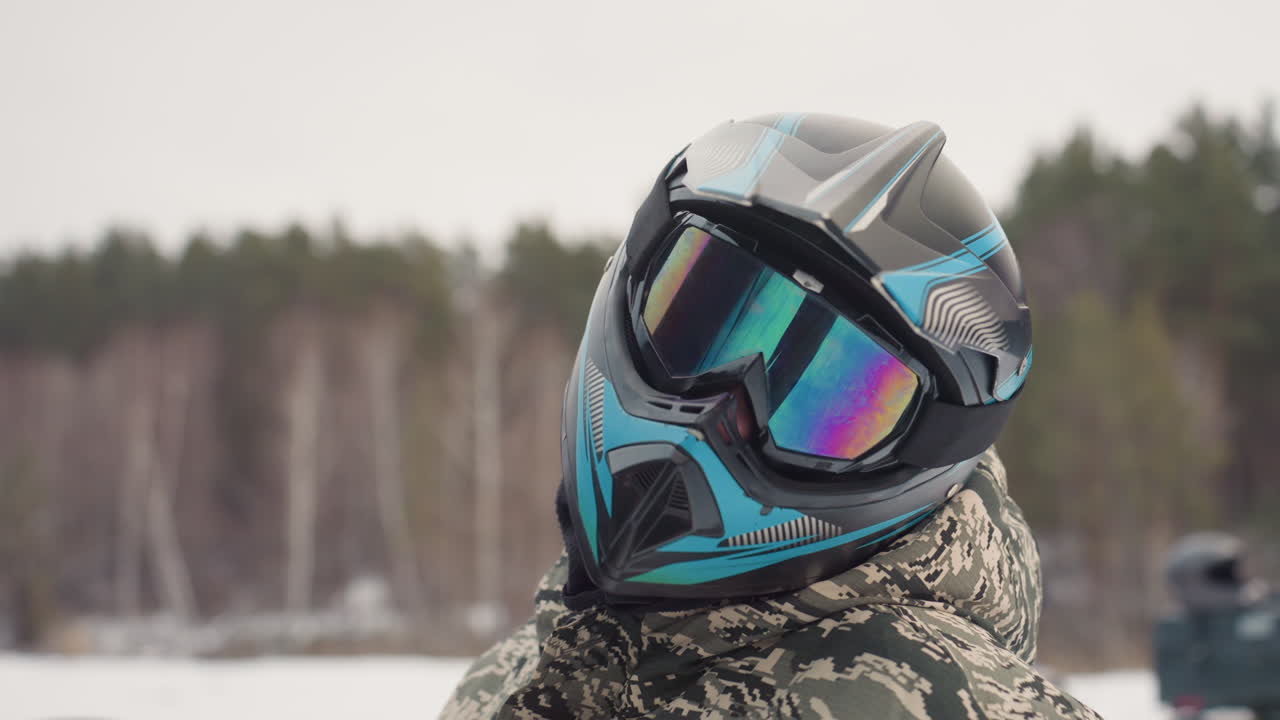 Atv rider with reflective helmet, camo jacket, snowy forest background, mirrored goggles catching overcast light, steady preride posture, checking visor and straps, calm anticipation, soft cold