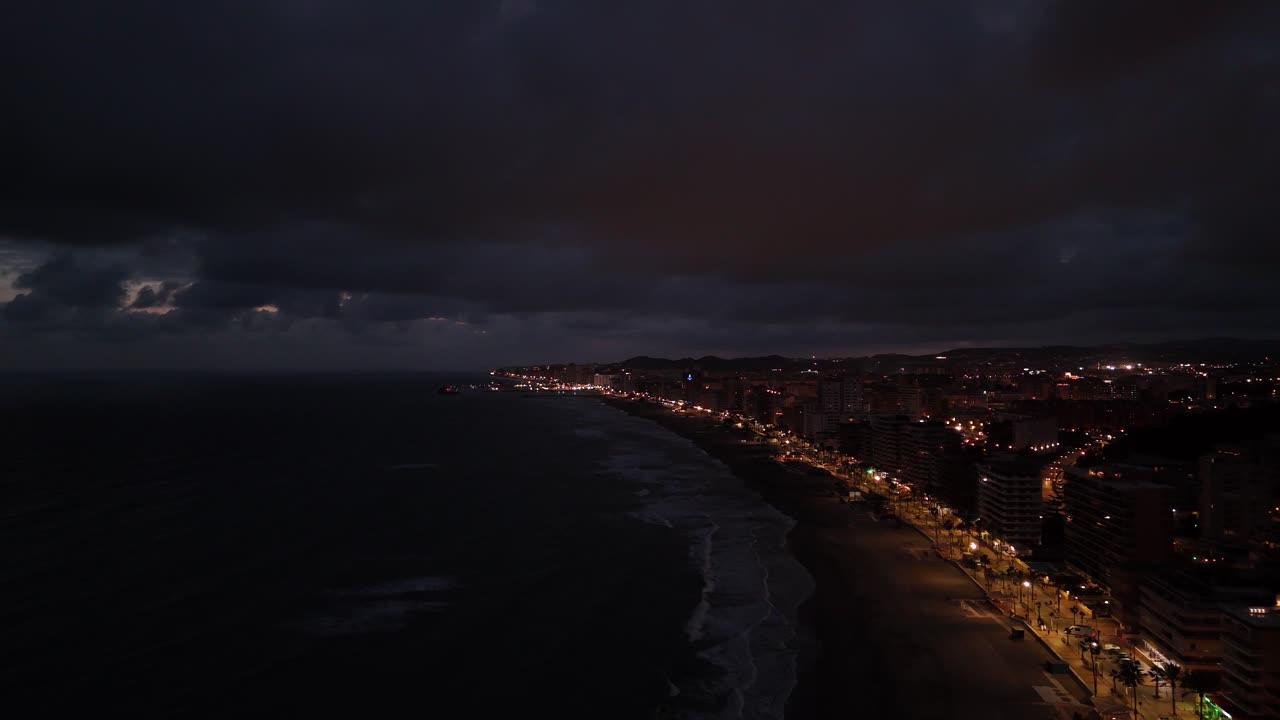 Aerial view of Torreblanca, Spain coastline at night showing illuminated buildings, dark sea, and cloudy sky, creating a dramatic and moody atmosphere, drone pulling out