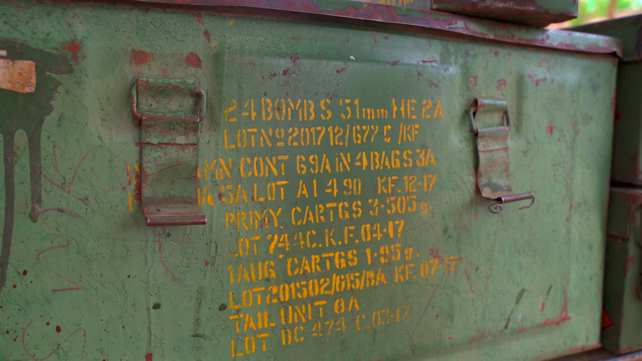 Una ripresa statica ravvicinata di una cassa di munizioni in metallo verde invecchiata con specifiche militari e numeri di lotto stampati in giallo a stencil, che mostra ruggine e usura autentiche
