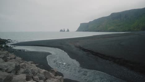 Der-Fluss-Fließt-Ins-Meer-Am-Schwarzen-Sandstrand-Von-Víkurfjara-In-Vik,-Island,-In-Der-Ferne-Reynisfjall