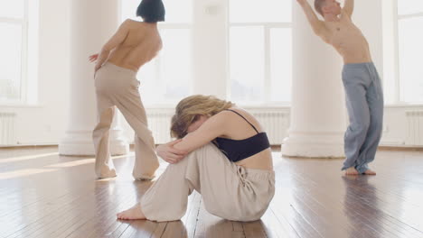 tänzerin, die mit dem kopf zwischen den beinen auf dem boden sitzt, während ihre kumpels im studio tanzen