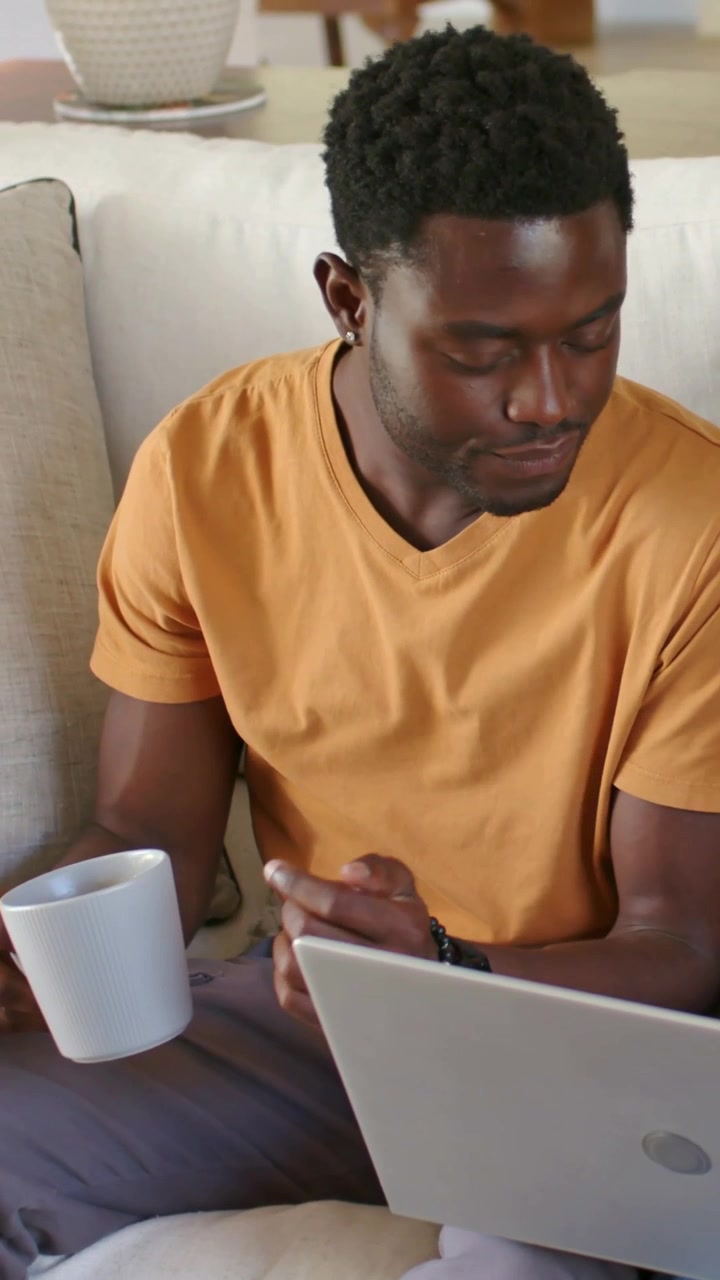 Vertical video: Working on laptop, man in mustard tee seated on couch tapping pad and holding mug