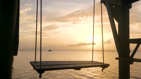 Un-Columpio-De-Madera-Vacío-Se-Balancea-En-Un-Muelle-Con-Vista-Al-Océano-Con-Un-Bote-En-Los-Rayos-Naranjas-De-La-Puesta-De-Sol