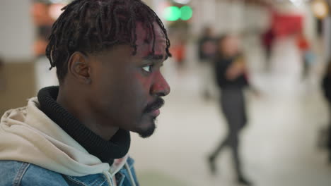 close-up shot of young person with short dreadlocks in denim jacket and hoodie, focused on interacting with digital screen in modern mall. blurred background with mall lights and people visible