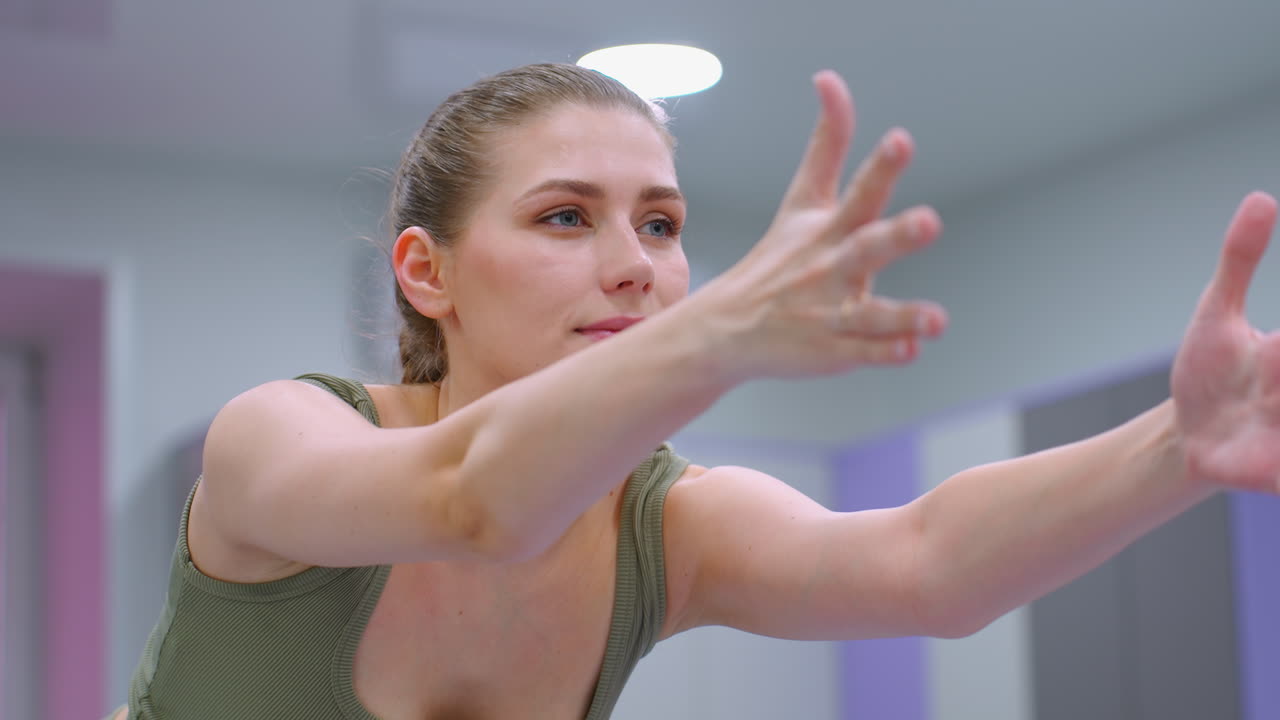 сосредоточенная молодая женщина выполняет приседание, протягивая руки вперед в ярко освещенном тренажерном зале, освещенном потолочными огнями, с решительным выражением лица и сильной позой